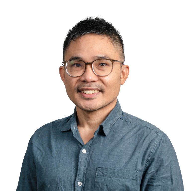 Smiling man with short black hair and brown-framed glasses, wearing a blue button-down shirt against a white background.