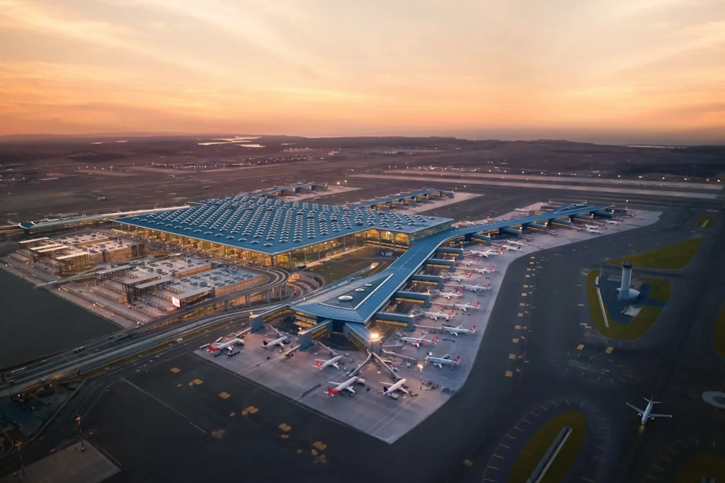 Aerial view of a large airport terminal at sunset, with concourses, many parked airplanes, and runways nearby under a orange-pink sky.