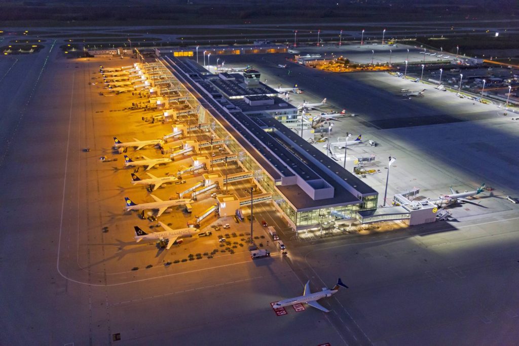Aerial night view of a busy airport terminal with numerous Lufthansa airplanes docked at gates.