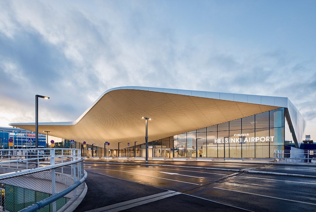Helsinki Airport terminal with a curved, overhanging roof and glass entrance at dusk/early evening.
