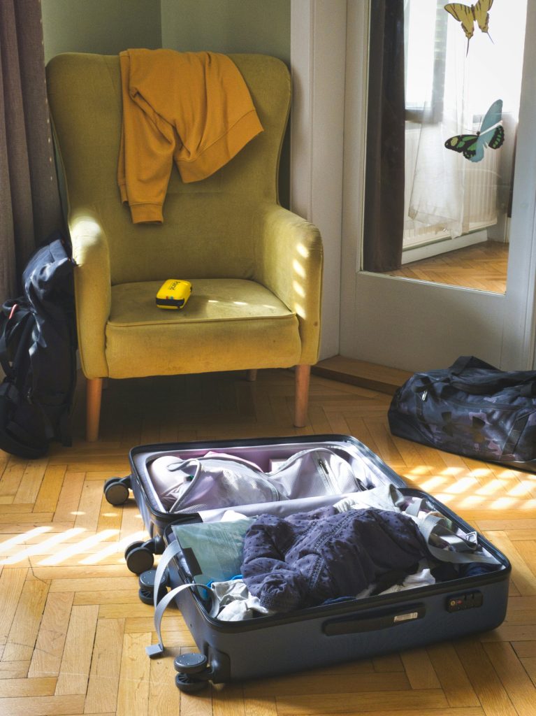 Yellow armchair with a mustard sweater draped over it, a small yellow tape measure on the seat, and an open suitcase full of clothes on a wooden floor by a glass door with butterfly decals.