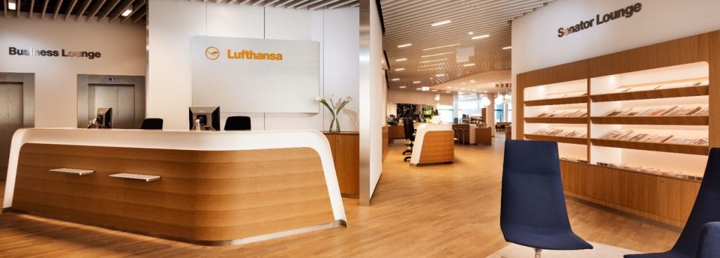 Wide shot of an airport business lounge with a curved wooden reception desk, Lufthansa signage, and a polished wooden floor. On the left are elevators labeled Business Lounge; on the right, shelves in a Senator Lounge area with blue chairs nearby.