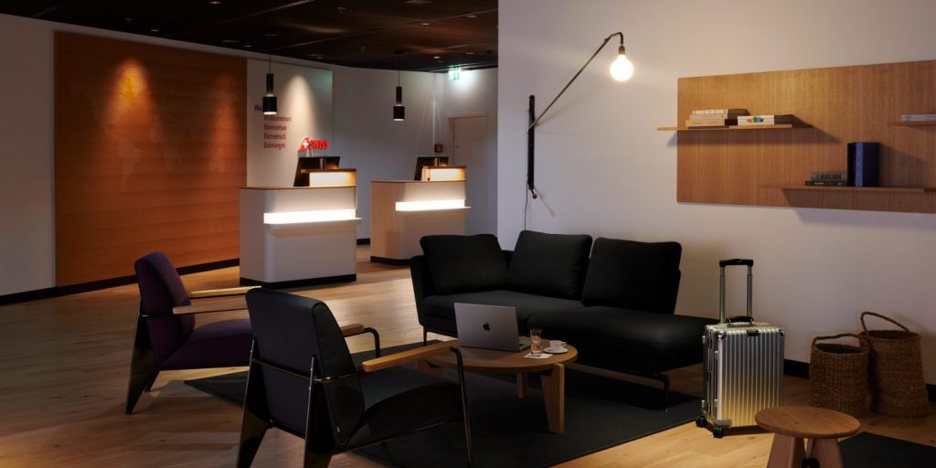 Modern hotel lobby with black sofa, wooden coffee table, laptop, and a silver suitcase near a wicker basket on the right.
