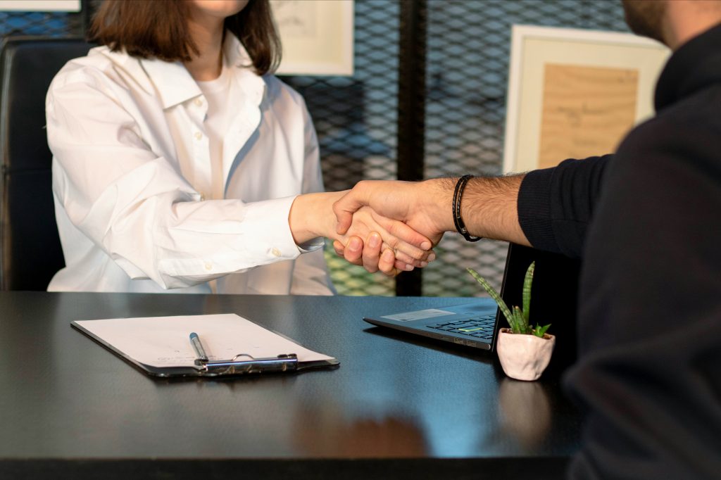 Two people shake hands across a desk in an office, with a notebook, pen, and laptop nearby and a small plant in the foreground