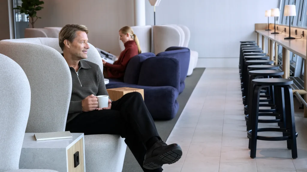 Man in a gray sweater sits in a curved chair, holding a mug, in a modern lounge foregrounding the scene.
