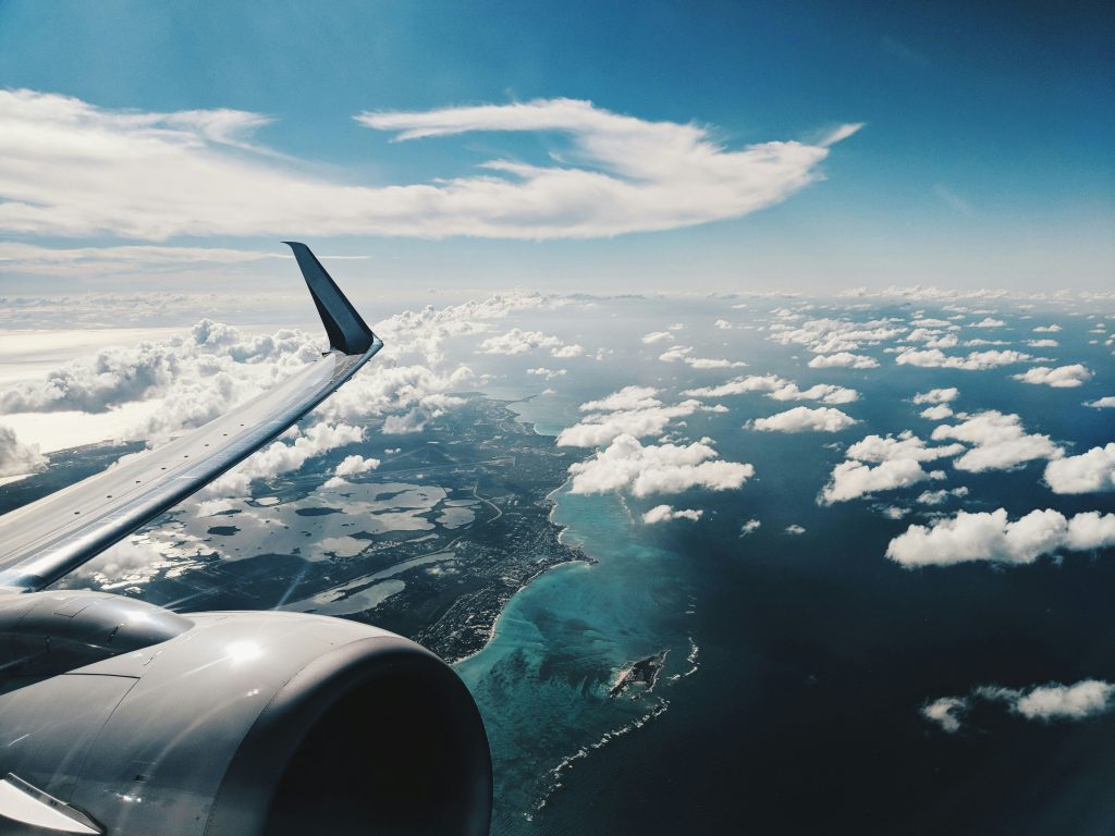 Airplane wing visible with ocean and cloud-covered coastline below, blue sky overhead.