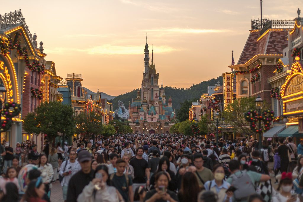 Crowded main street at dusk with festive lights and a distant castle in a fantasy theme park.