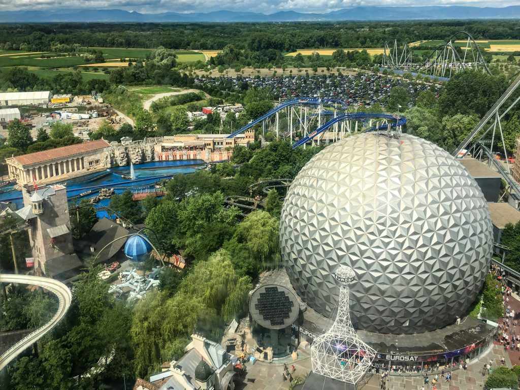 Aerial view of a theme park with a large geodesic dome, blue roller coasters, trees, and a distant mountain backdrop.