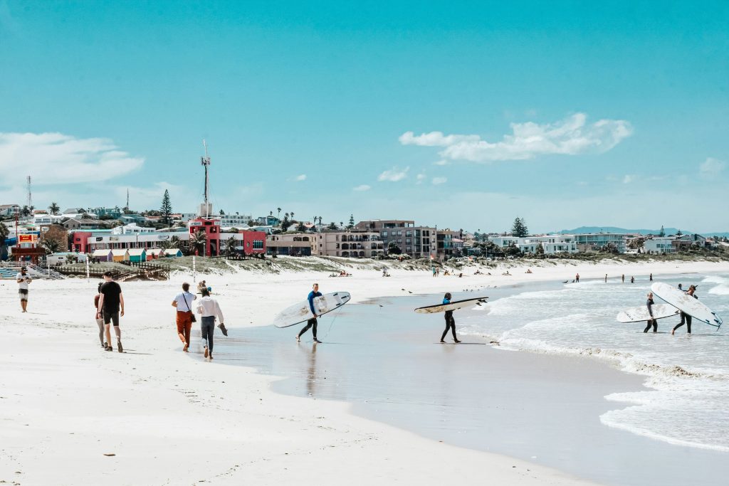People carrying surfboards and walking along a sunny beach with a town skyline and blue sky in the background.