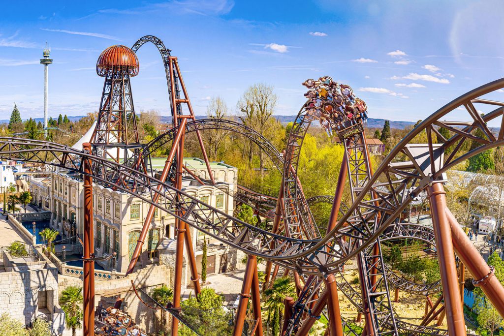 Aerial view of a steel roller coaster with riders looping and twisting through brown tracks under a blue sky.