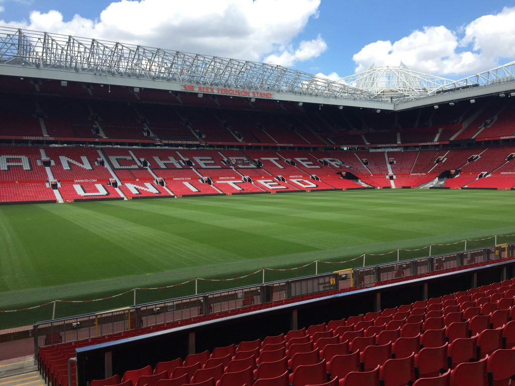 Football stadium with red seating spelling 'MANCHESTER UNITED' along the stands and a green pitch beneath a blue sky, view from near the field's edge.