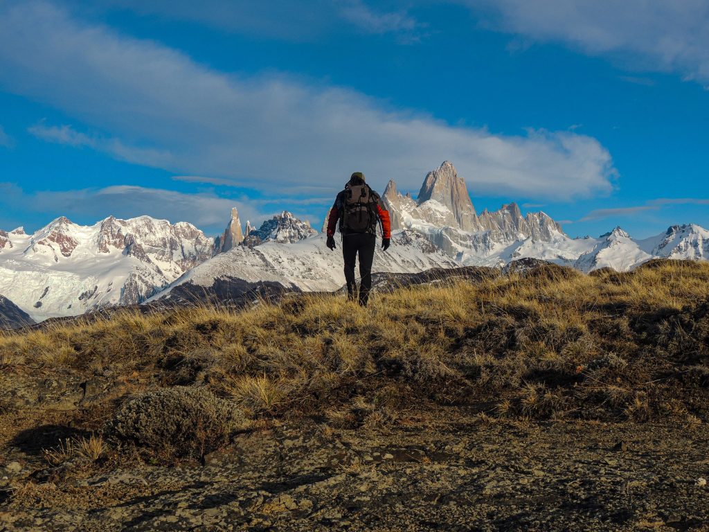 Hiker with backpack standing on a grassy ridge, facing snow-capped mountains under a blue sky.