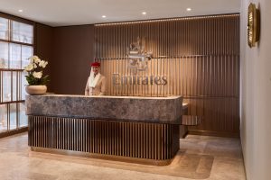 Reception desk at Emirates counter with smiling attendant in beige uniform and red hat, marble counter and bronze slat wall backdrop showing Emirates logo.