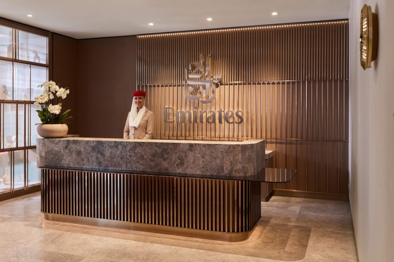 Reception desk at Emirates counter with smiling attendant in beige uniform and red hat, marble counter and bronze slat wall backdrop showing Emirates logo.