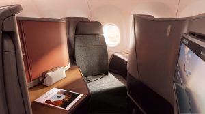 Business class airplane seat with a high back, window view, personal screen to the right, and a travel magazine on a wood-toned desk.