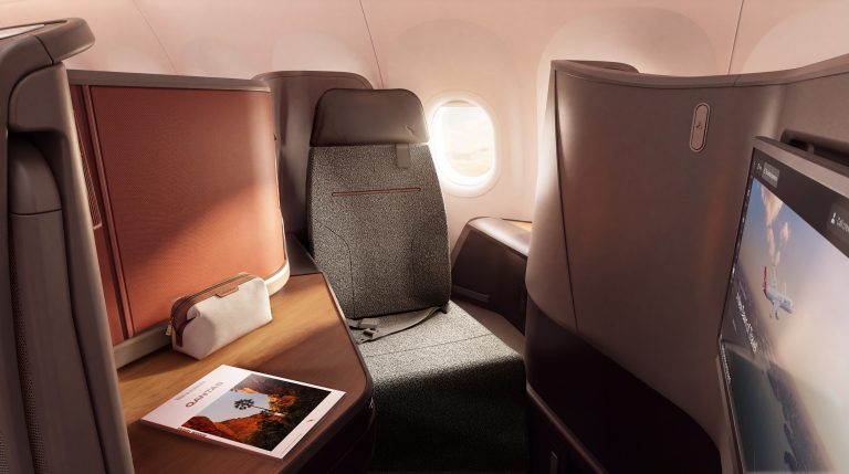 Business class airplane seat with a high back, window view, personal screen to the right, and a travel magazine on a wood-toned desk.