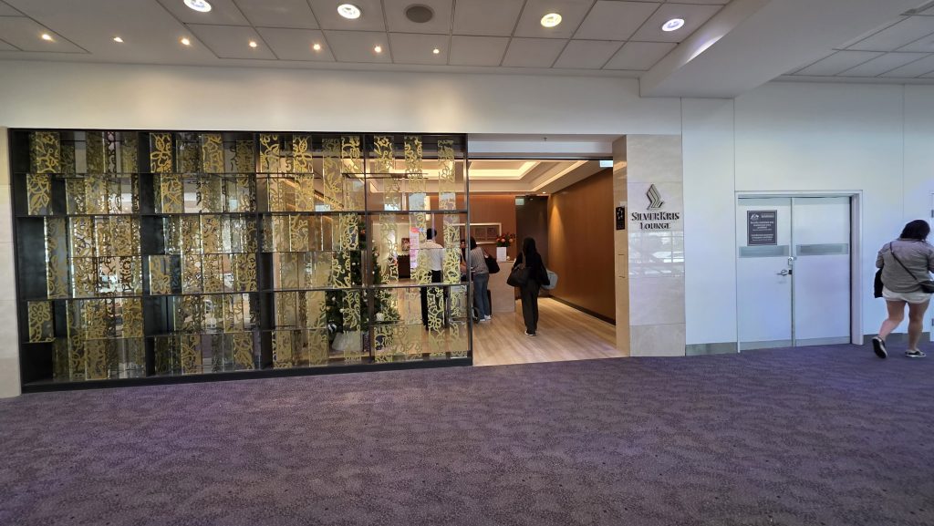 Entrance to a lounge area with decorative glass panels featuring a gold floral pattern; people stand at the doorway and a sign on the wall reads the lounge name.